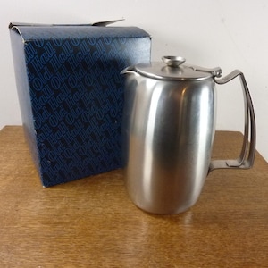 May include: A stainless steel coffee pot with a lid and handle, next to a blue box with the text "Hall" repeated. The pot has a curved body and a spout. The box has a dark blue background.