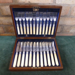 Puede incluir: Una caja de madera que contiene un juego de cubiertos de plata con mangos de marfil. La caja está abierta y los cubiertos están dispuestos en dos filas. Los cubiertos incluyen cuchillos, tenedores y cucharas.