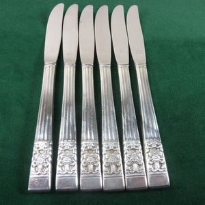 Puede incluir: Seis cuchillos plateados con un patrón floral decorativo en los mangos. Las hojas son curvas y los mangos tienen estrías verticales. Los cuchillos están colocados sobre una superficie verde.