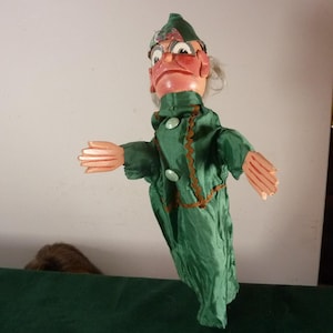May include: A hand puppet featuring a character with a painted face, glasses, and a green hat. The puppet wears a long-sleeved green satin-like robe with two light green buttons and brown decorative trim along the front and sleeves.