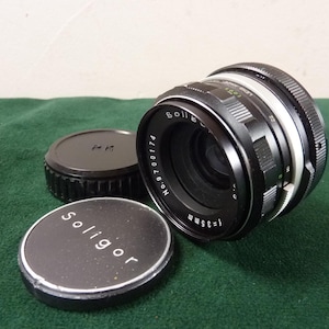 Puede incluir: Un objetivo de cámara Soligor negro con un anillo de ajuste blanco y plateado, y dos tapas de objetivo negras. El objetivo está marcado con "f=35mm" y "Soligor". Las tapas del objetivo son negras con texto blanco.