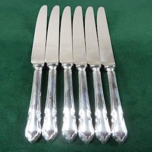 May include: Six silver knives with decorative handles are displayed on a green surface. The blades are smooth and shiny, and the handles have an ornate design. These knives are likely for use at a dining table.