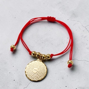 May include: Red string bracelet with gold-colored beads and a circular pendant. The pendant has etched text and is suspended from the red string. The bracelet is adjustable.