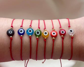 May include: A collection of seven red string bracelets, each featuring a glass evil eye charm in a different color. The charms are round with a white iris and black pupil, and are accented with gold beads.