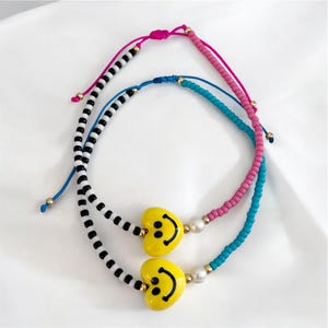 Puede incluir: Dos pulseras de cuentas. Una presenta cuentas a rayas blancas y negras, la otra tiene cuentas rosas y turquesas. Ambas tienen dijes amarillos en forma de corazón con caras sonrientes. Las pulseras son ajustables con cordones rosas y azules.