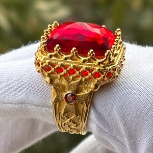 May include: A gold-colored ring featuring a large, faceted red gemstone. The ring has an ornate design with smaller red accents and intricate detailing. The ring is held by a gloved hand against a blurred green background.