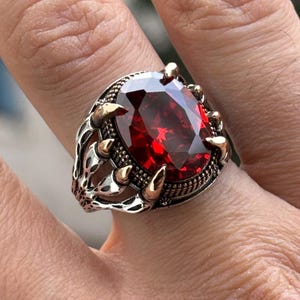 Puede incluir: Anillo de plata con una gran gema roja oscura de corte ovalado. El anillo tiene un intrincado trabajo de metal con detalles dorados alrededor de la piedra y en la banda. El diseño presenta garras que sujetan la gema.