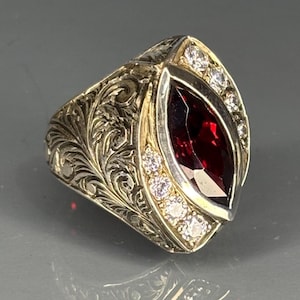 Könnte beinhalten: Ein silberner Ring mit einem großen, marquiseförmigen, tiefroten Edelstein. Der Ring ist mit kleineren, klaren Edelsteinen und aufwendigen floralen Gravuren auf dem Band verziert. Die Fassung ist goldfarben und erzeugt einen auffälligen Kontrast.