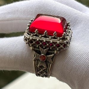 May include: A silver ring featuring a large, square, red gemstone. The ring has an ornate, detailed setting with smaller red stones and a cross design on the band. The ring is shown on a white surface.