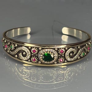 May include: Silver cuff bracelet with gold accents, featuring an oval green gemstone at the center, surrounded by smaller red and green gemstones. The bracelet has an ornate design with swirling patterns and clear, sparkling accents.