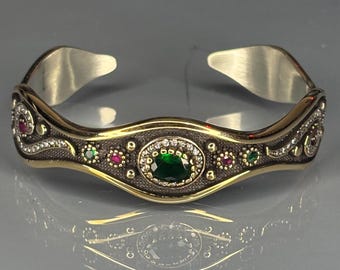 Pode incluir: Pulseira aberta com uma faixa prateada e um centro decorativo dourado. O centro apresenta uma pedra preciosa verde oval rodeada por pedras brancas, com pedras preciosas vermelhas e verdes menores. A pulseira é exibida em uma superfície reflexiva.