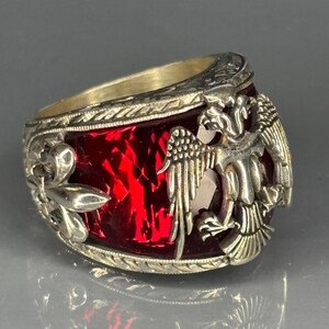 Close-up view of a heraldic men's ring showcasing a fiery faceted ruby gemstone set in 925 sterling silver. The sides feature a double-headed eagle motif, symbolizing strength and unity. The silver scrollwork and the vibrant red gem give this piece a bold, medieval-inspired look.