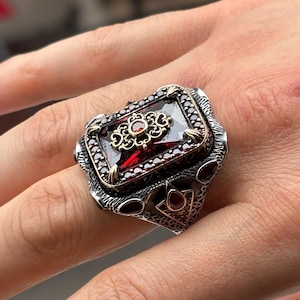 May include: A silver ring with a rectangular, deep red gemstone at the center. The ring features intricate gold and silver detailing around the stone and on the band, with smaller red stones accenting the design. The ring is worn on a finger.