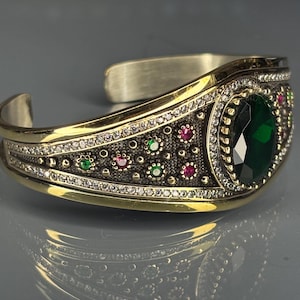 May include: A gold and silver cuff bracelet with an oval, faceted, emerald-green gemstone as the focal point. The bracelet is adorned with small, round, red, green, and clear gemstones. The design features intricate detailing and a vintage aesthetic.