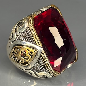 May include: A silver ring with gold detailing and a large, rectangular, red gemstone. The ring features intricate patterns on the band and around the gemstone. The red gemstone is faceted, reflecting light and adding to its brilliance.