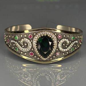 Puede incluir: Una pulsera de puño dorada con un diseño ornamentado. La pulsera presenta una gran piedra preciosa verde oscuro en forma de pera en el centro, rodeada de piedras preciosas rojas, verdes y transparentes más pequeñas. La banda tiene un patrón intrincado y ondulado.