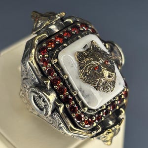 May include: A detailed silver ring featuring a bear's head with red gemstone eyes set on a white stone. The ring is accented with red gemstones and gold-colored details, showcasing intricate craftsmanship.