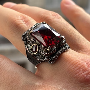 Puede incluir: Un anillo de plata con una gran piedra preciosa rectangular de color rojo intenso. El anillo tiene un diseño ornamentado con piedras más pequeñas de color rojo oscuro y detalles dorados. El anillo está en una mano.