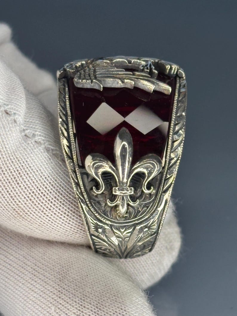 May include: A silver ring with a rectangular, deep red stone inset with two white diamond shapes. The ring features a fleur-de-lis design and intricate floral engravings. The top of the ring has a detailed crest.