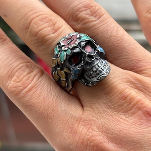 Op de afbeelding: Een zilveren schedelring met kleurrijke bloemaccenten. De ring heeft een gedetailleerd schedelontwerp met rode, roze, blauwe en groene emaille. De bloemen zijn ingewikkeld gedetailleerd en zorgen voor een levendig contrast.