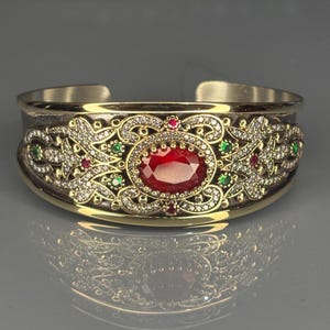 May include: A gold and silver cuff bracelet with a central oval, red gemstone. The bracelet has intricate filigree and is decorated with small, sparkling white, green, and red gemstones. The design is symmetrical and detailed.