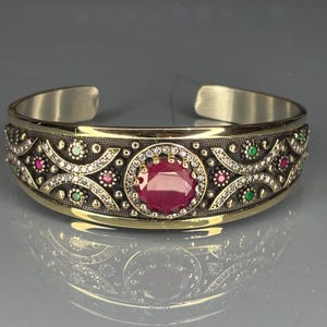 Puede incluir: Un brazalete de plata y oro con una gran gema roja redonda en el centro. El brazalete está adornado con piedras preciosas más pequeñas en verde, rosa y blanco, y detalles de filigrana intrincados.