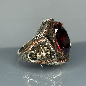 May include: A silver ring with an ornate design and a large, dark red gemstone. The ring has a small loop on the side.
