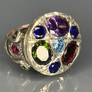 Puede incluir: Un anillo de plata con un diseño circular con múltiples piedras preciosas. El anillo presenta una variedad de colores, incluyendo piedras preciosas moradas, azules, verdes, rojas y celeste. El anillo tiene una banda ornamentada y detallada.