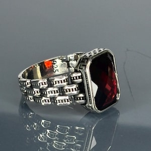 May include: A silver ring with a red gemstone and a chain-like band. The ring is marked with "925" on the inside.