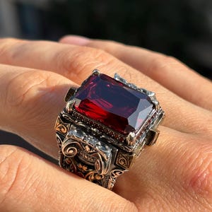 Puede incluir: Un anillo ornamentado con una gran gema rectangular de color rojo intenso. El anillo tiene una banda detallada en tono plateado con detalles dorados y un intrincado trabajo de volutas. La gema es el punto focal, captando la luz y añadiendo un toque de lujo.