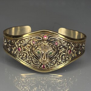 May include: A gold-coloured cuff bracelet with an ornate design, featuring a jaguar head at the centre. The bracelet is adorned with red and green gemstones. It has an open design for easy wear.