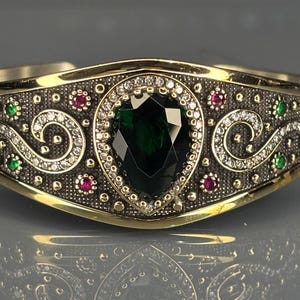 Könnte beinhalten: Ein goldfarbenes Armreif-Armband mit einem großen, birnenförmigen, dunkelgrünen Edelstein in der Mitte. Das Armband weist aufwendige Details mit kleinen roten, grünen und weißen Edelsteinen sowie ein dekoratives Muster auf.