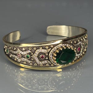 May include: A gold-coloured cuff bracelet with an intricate pattern. The bracelet has a large, oval, emerald-green gemstone at the centre, surrounded by smaller red and green stones and clear crystals. The bracelet has an open design.