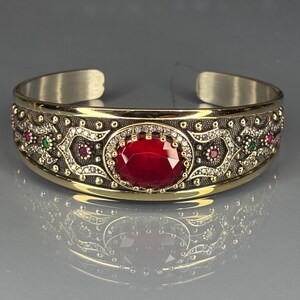 Puede incluir: Una pulsera de puño dorada y plateada con una gran gema roja ovalada en el centro. La pulsera presenta detalles intrincados con pequeñas gemas rojas, verdes y blancas, y un patrón decorativo a lo largo de la banda.