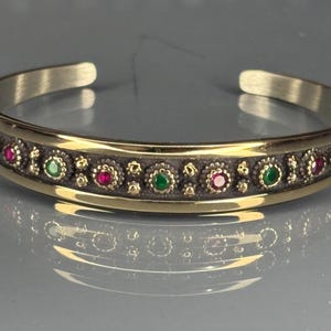 May include: A gold-coloured cuff bracelet with a dark band featuring alternating red and green gemstones. The bracelet has a detailed, ornate design with a polished finish. The overall style is elegant and decorative.