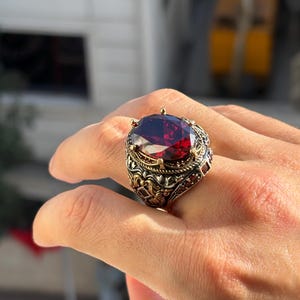 Può includere: Un anello ornato con una grande gemma ovale rosso scuro. L'anello presenta un design a filigrana color oro e argento antico attorno alla pietra e alla fascia. La gemma è il punto focale.