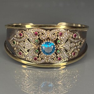 May include: An ornate cuff bracelet with silver and gold detailing. The bracelet features a large, round, blue gemstone at the centre, surrounded by smaller red, green, and clear gemstones. The design includes intricate filigree and scrollwork.