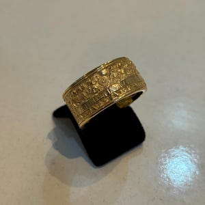 Pode incluir: Um anel de ouro com detalhes intrincados e uma banda larga. O anel está colocado em um expositor de veludo preto.