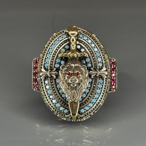 Può includere: Un anello d'argento con un designo di pietre blu e rosa. L'anello presenta una testa di leone con occhi rossi e una spada.