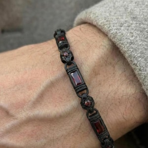 May include: A black bracelet with rectangular and round red gemstones. The bracelet is made of a dark metal and features an ornate design. The gemstones are a deep red color, and the bracelet is worn on a wrist.