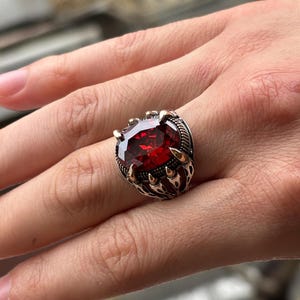 May include: A silver ring with a large, oval-cut, deep red gemstone. The ring has a detailed, claw-like setting with gold-colored accents. The ring is on a person's hand, showcasing the jewelry's design and color.