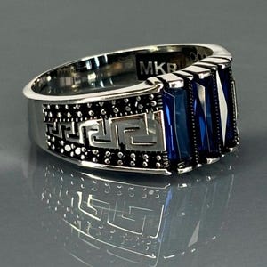 May include: A silver ring featuring a blue stone and a Greek key design. The ring is marked "MKR" on the inside.