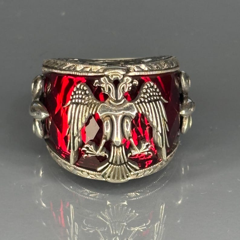 Side-angle image of the ruby ring highlighting its wide band and raised gemstone setting. The intricate details of the double-headed eagle emblem are visible on the shoulder, while the silver has a natural, uncoated finish. The ruby’s red tone glows brightly against the sculpted silver.