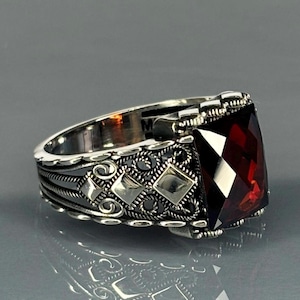 Könnte beinhalten: Ein silberner Ring mit einem großen, dunkelroten Edelstein, der in einem filigranen Design gefasst ist. Der Ring hat ein detailliertes, kompliziertes Design mit kleinen silbernen Akzenten.