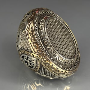 May include: Ornate silver ring with gold accents. The ring features intricate filigree and a textured oval center. The design includes scrolling patterns and geometric shapes, creating a detailed and luxurious appearance. The ring is a statement piece.