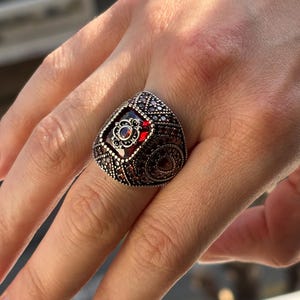 May include: A detailed, ornate ring with a large, rectangular red gemstone at its center. The ring is dark silver with intricate patterns and smaller red stones. The design is symmetrical and features a floral-like motif.