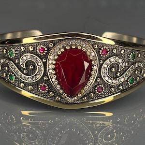 May include: A gold-toned cuff bracelet with a large, teardrop-shaped, red gemstone at its centre. The bracelet features intricate detailing with small red, green, and clear gemstones, and a decorative pattern. The design is symmetrical and ornate.