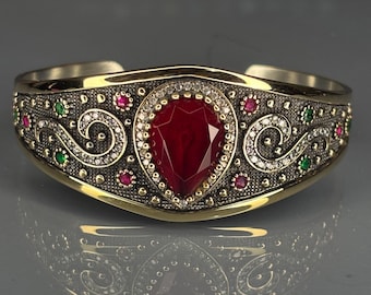 Pode incluir: Uma pulseira de punho dourada com uma grande pedra preciosa vermelha em forma de gota no centro. A pulseira apresenta detalhes intrincados com pequenas pedras preciosas vermelhas, verdes e transparentes e um padrão decorativo. O design é simétrico e ornamentado.