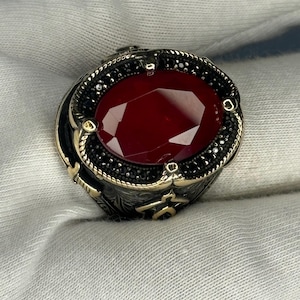 Può includere: Un anello d'argento con una grande pietra rossa ovale circondata da pietre nere. L'anello presenta dettagli intricati e una fascia testurizzata.