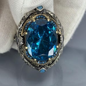Gloved close-up front view of the oval aquamarine blue stone set in a 925 sterling silver Ottoman-style men’s ring on gray background.